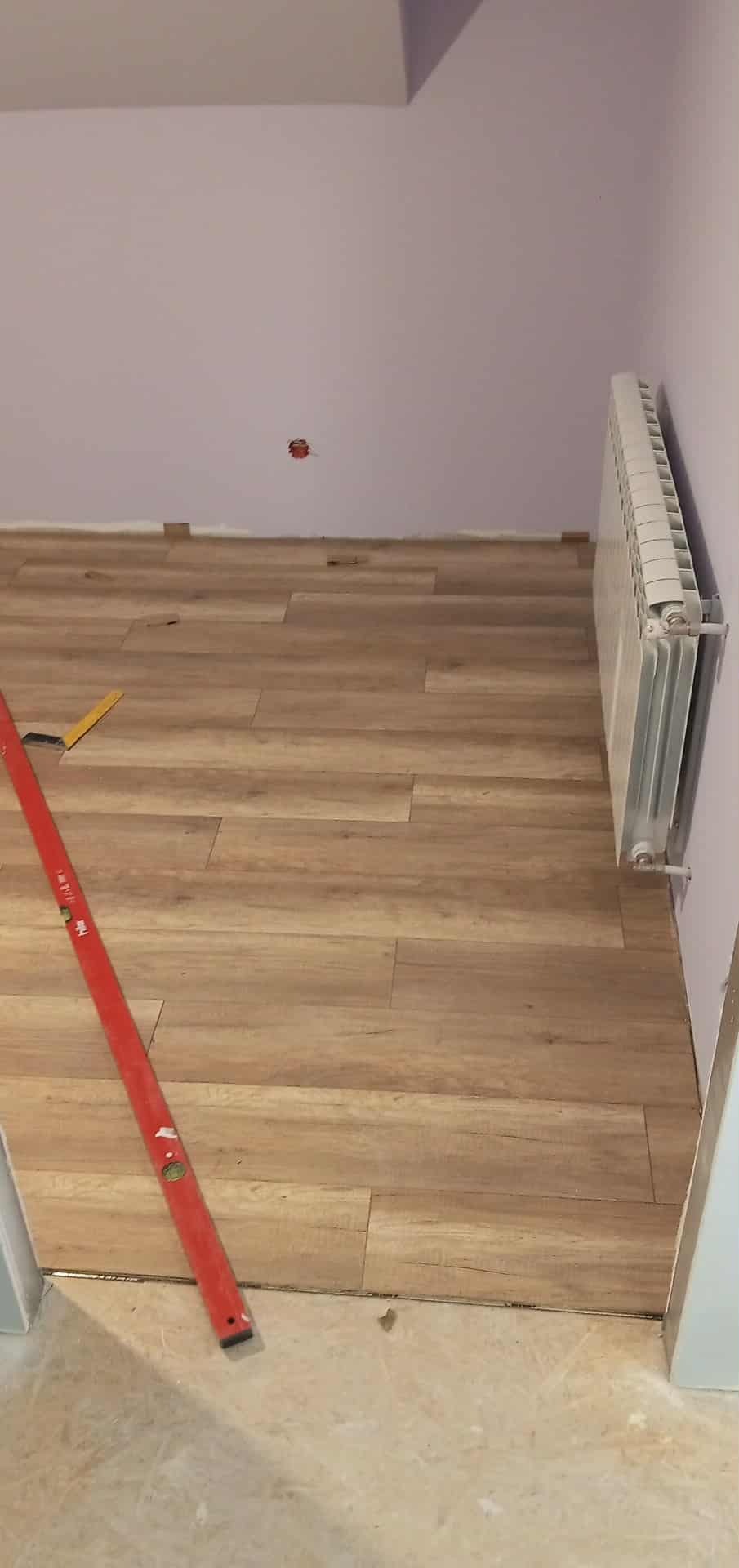 Fragment pokoju z ułożonymi panelami podłogowymi o wzorze drewna. Przy ścianie grzejnik, widoczna poziomica. Ściany w odcieniach fioletu. Częściowo widoczne podłoże przed panelami.