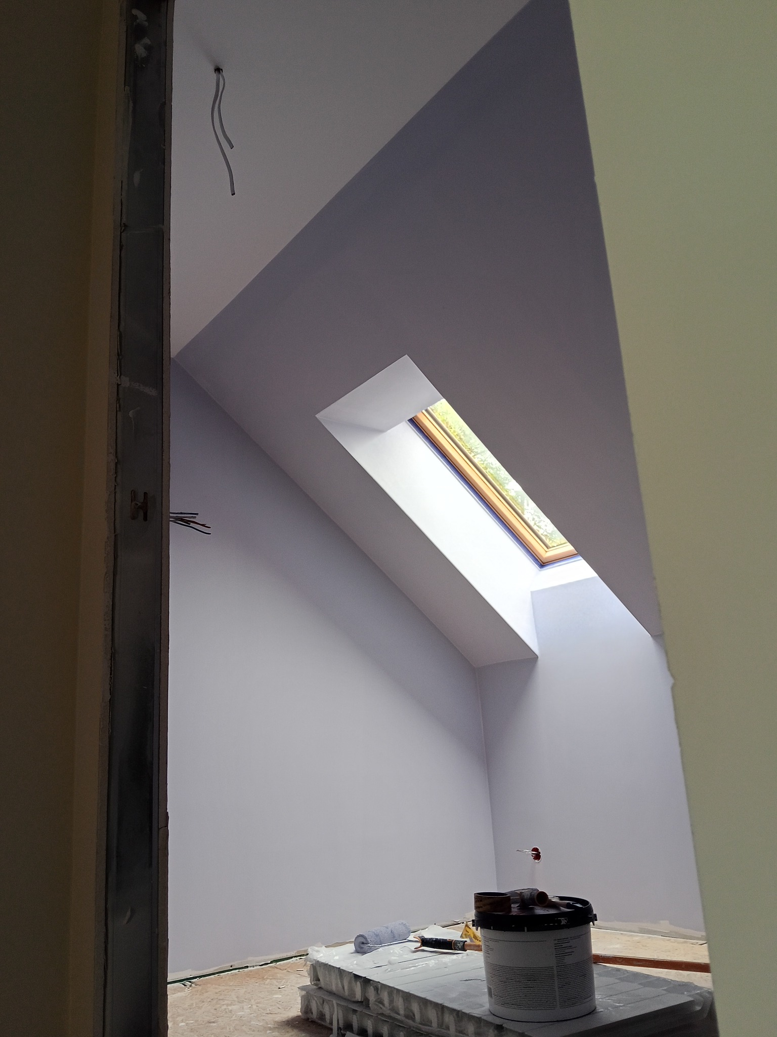 Pomalowane na szaro poddasze z oknem dachowym, widok zza framugi. Na podłodze wiadro z farbą i wałek malarski. Widoczne kable wystające z sufitu.