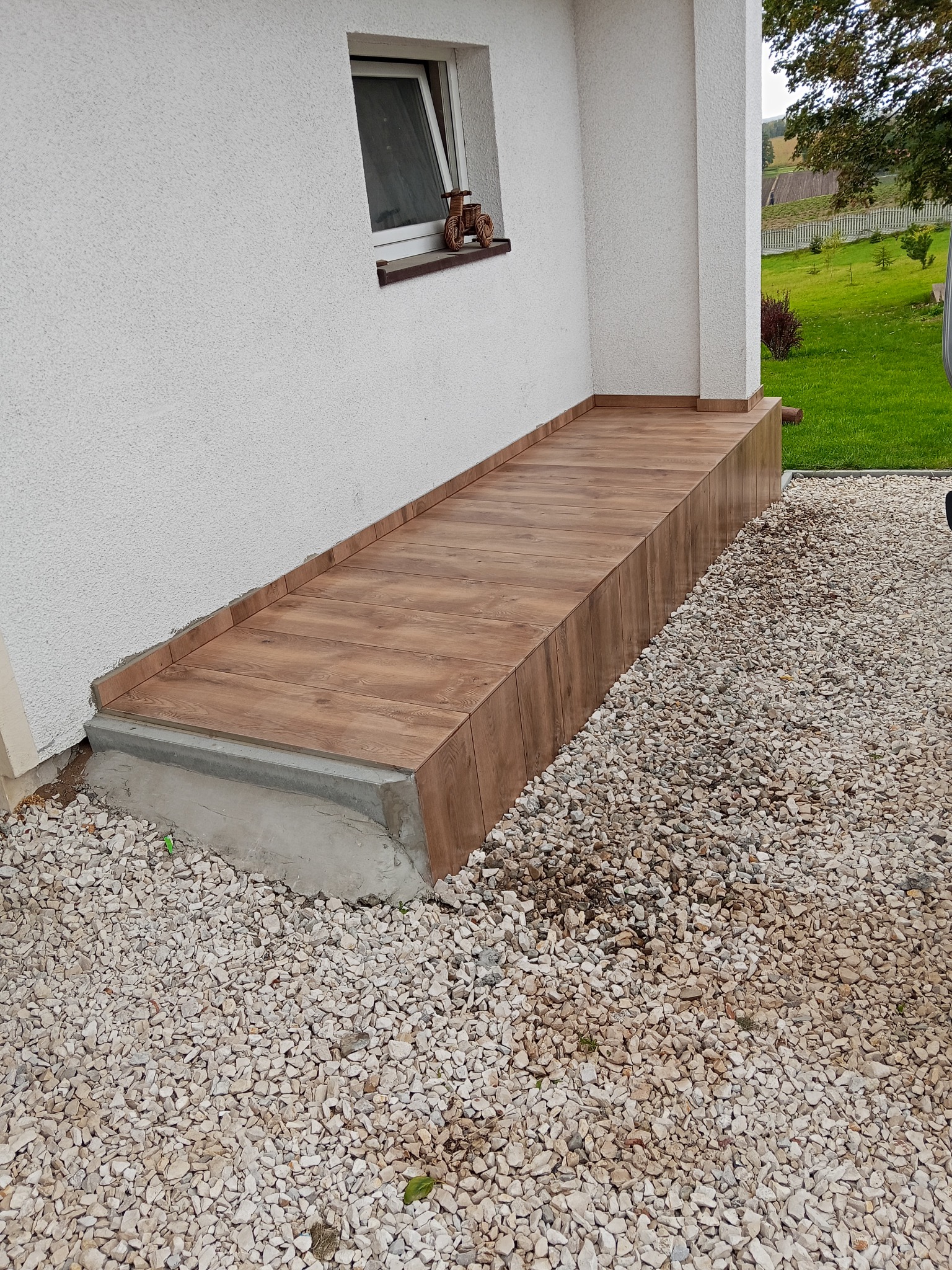 Wykończony taras z płytek imitujących drewno, przylegający do białej ściany budynku. Widoczne okno z dekoracją i żwirowe podłoże.