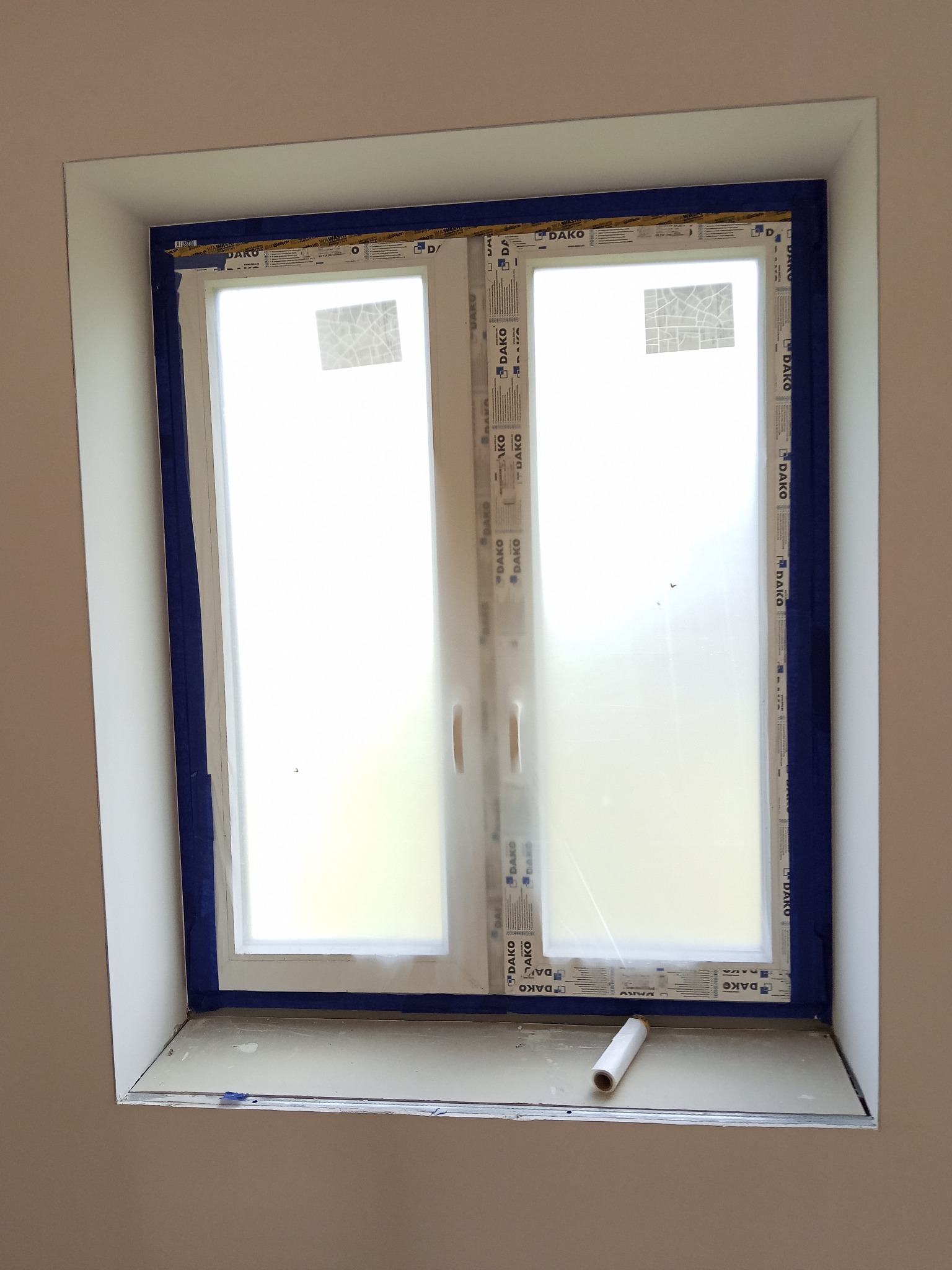 Okno DAKO zabezpieczone niebieską taśmą malarską przed malowaniem. Na parapecie leży rolka folii ochronnej. Przygotowanie do prac wykończeniowych.