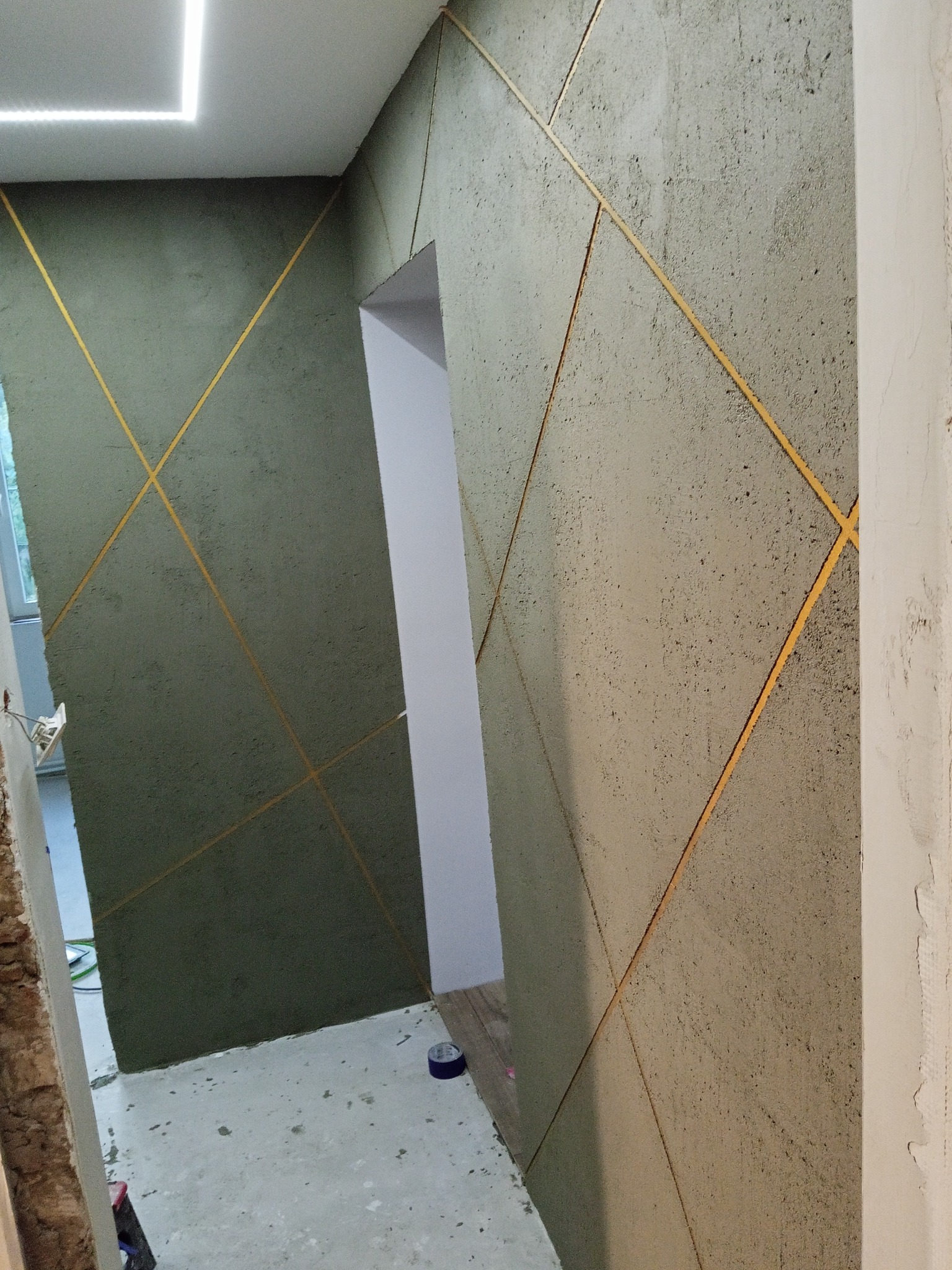 Dekoracyjna ściana z szorstką fakturą i złotymi liniami tworzącymi geometryczny wzór. Widoczne przejście do innego pomieszczenia i fragment sufitu z oświetleniem LED.