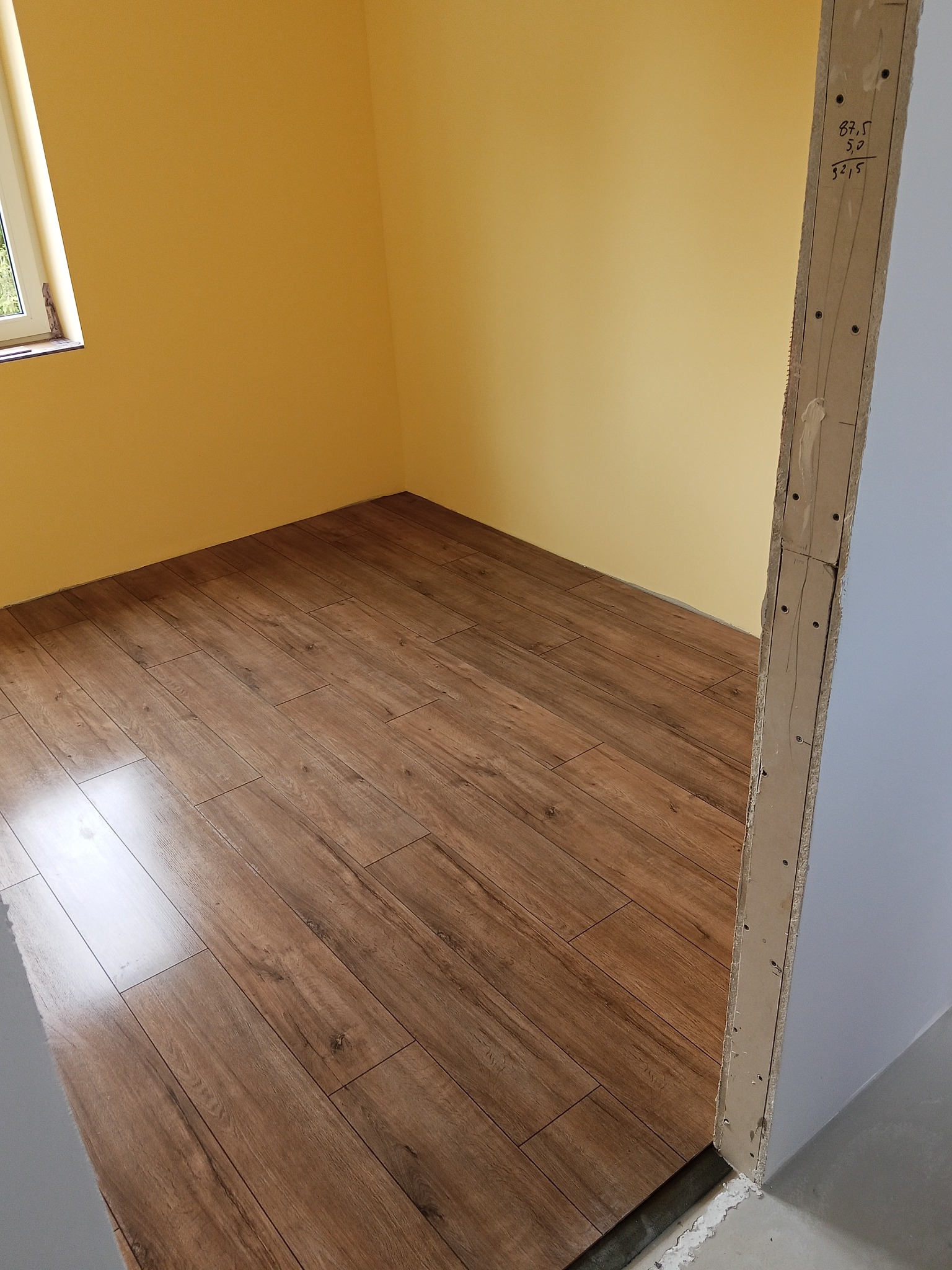 Ukończone układanie paneli podłogowych w pokoju z żółtymi ścianami. Widoczny fragment okna i surowa ościeżnica drzwi z naniesionymi notatkami.