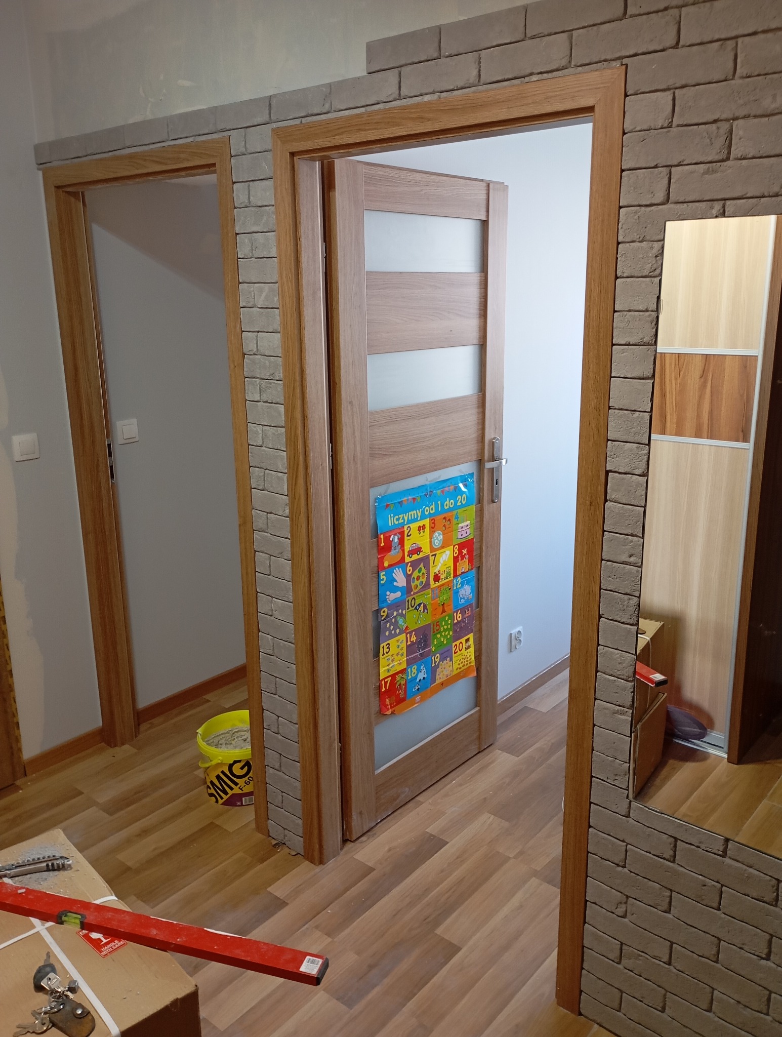 Wykończone ościeżnice drzwiowe z dekoracyjną cegłą, widoczne narzędzia i wiadro z zaprawą. Układanie paneli podłogowych.