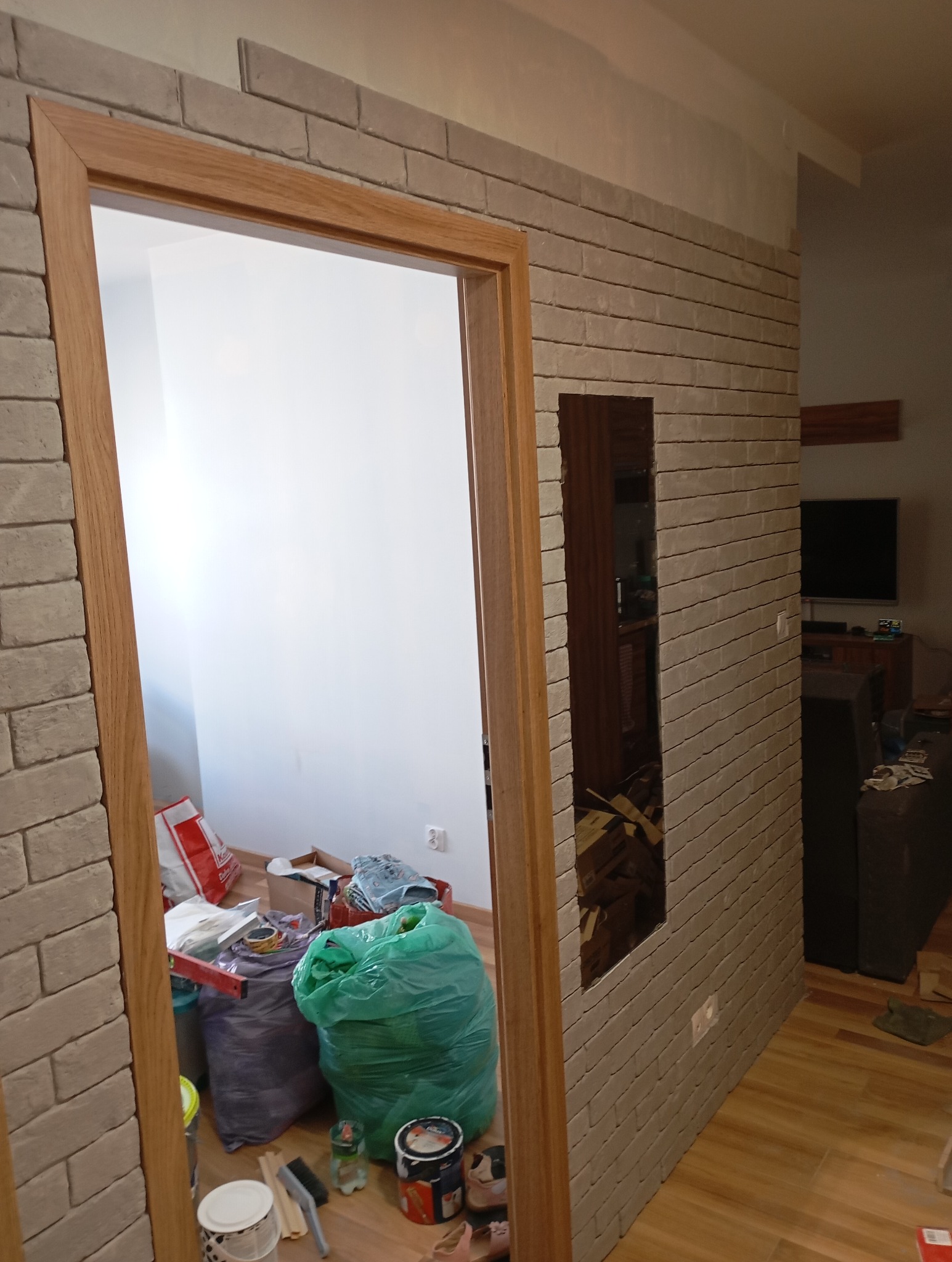 Ściana wykończona cegłą dekoracyjną z wnęką i drewnianą ościeżnicą. W tle widoczne worki z odpadami i narzędzia malarskie, co sugeruje etap prac wykończeniowych.