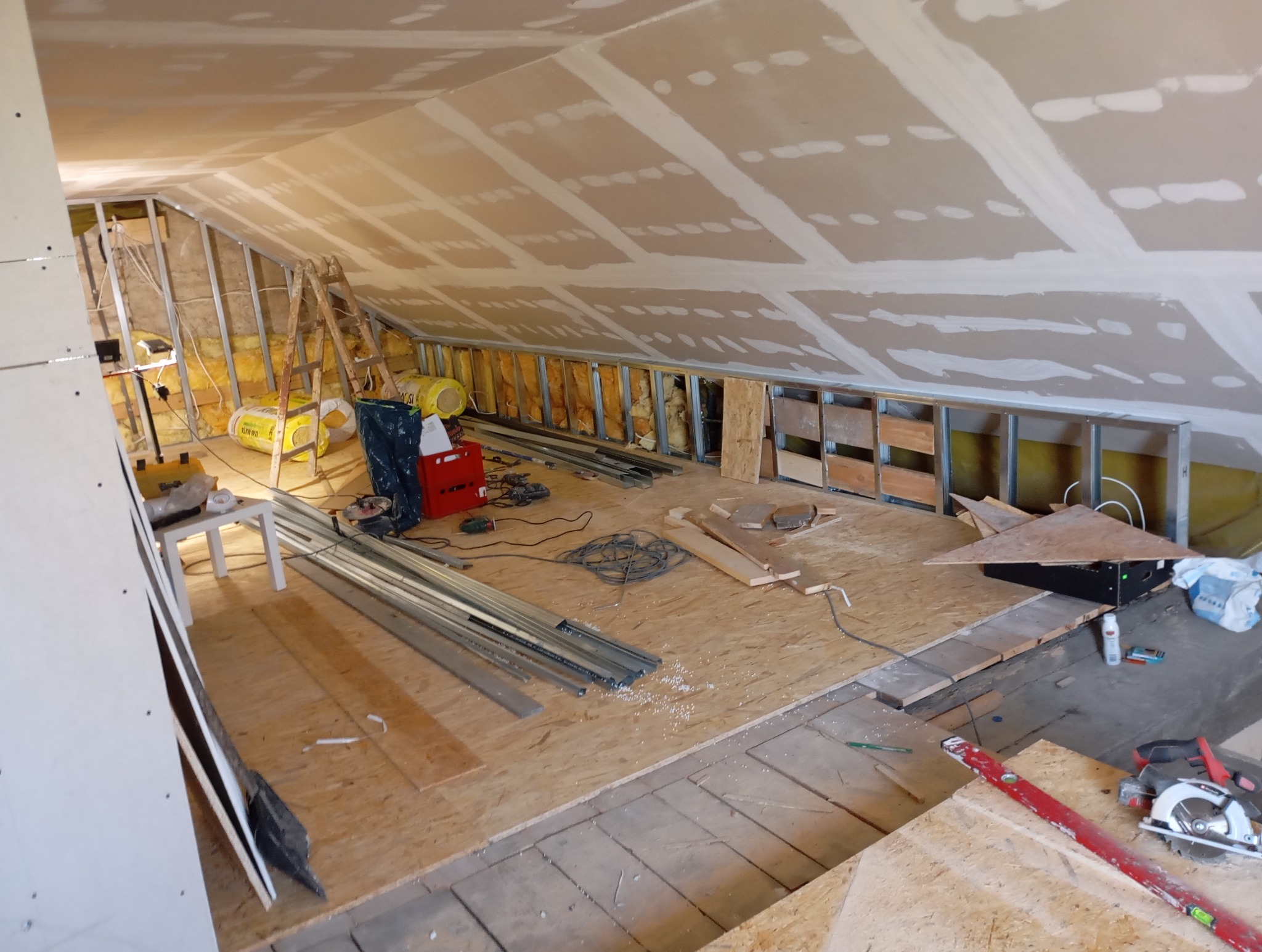 Poddasze w trakcie adaptacji: widoczne ocieplenie, stelaż z profili, płyty OSB na podłodze, narzędzia i materiały budowlane. Prace wykończeniowe w toku.