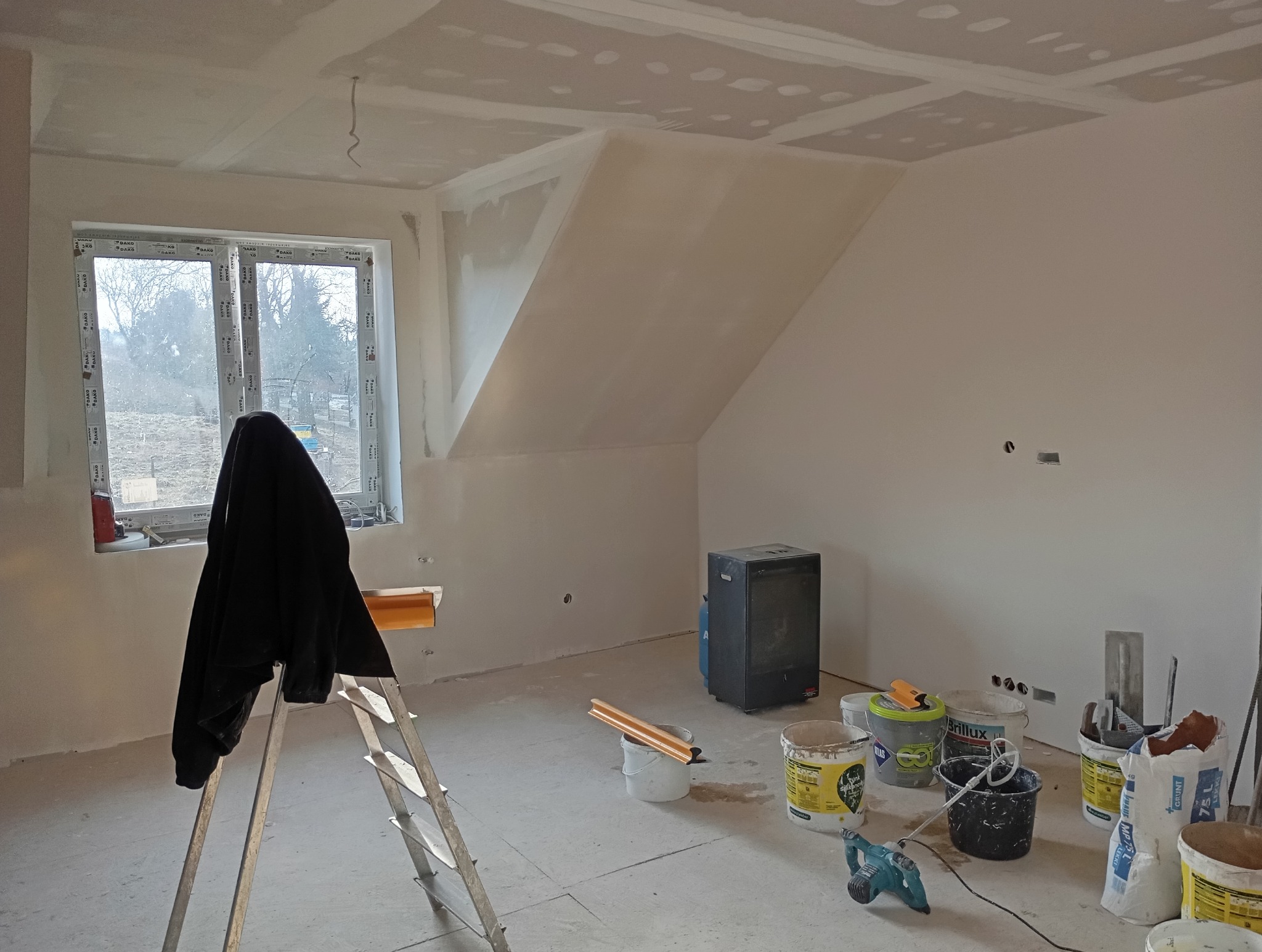 Poddasze po nałożeniu gładzi, widoczne narzędzia i drabina. Ściany i sufit przygotowane do malowania. Okno z widokiem na zewnątrz. Grzejnik elektryczny w rogu.