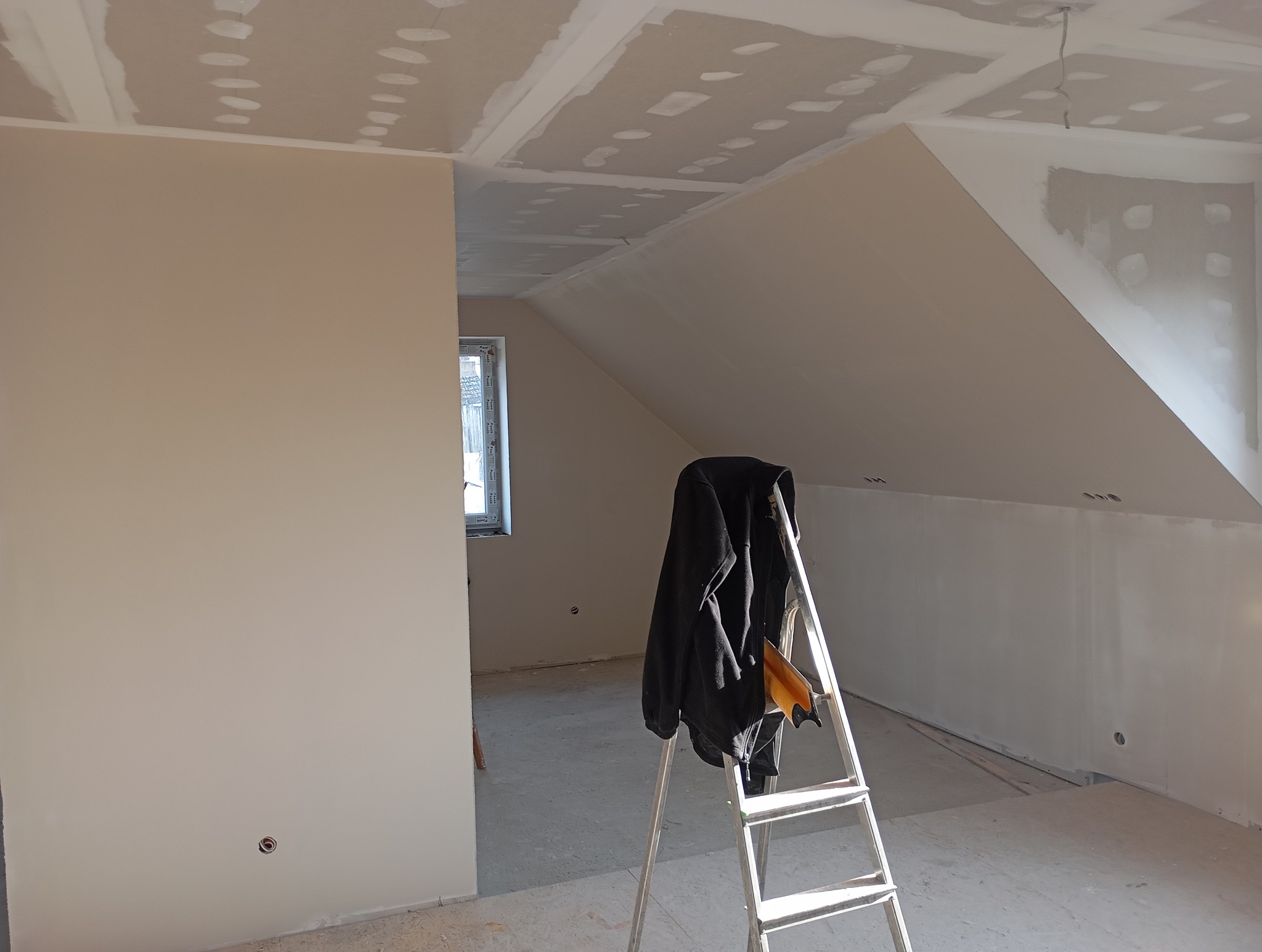 Poddasze po montażu płyt gipsowo-kartonowych, widoczna drabina z kurtką, przygotowane do dalszych prac wykończeniowych, jasne wnętrze z oknem.