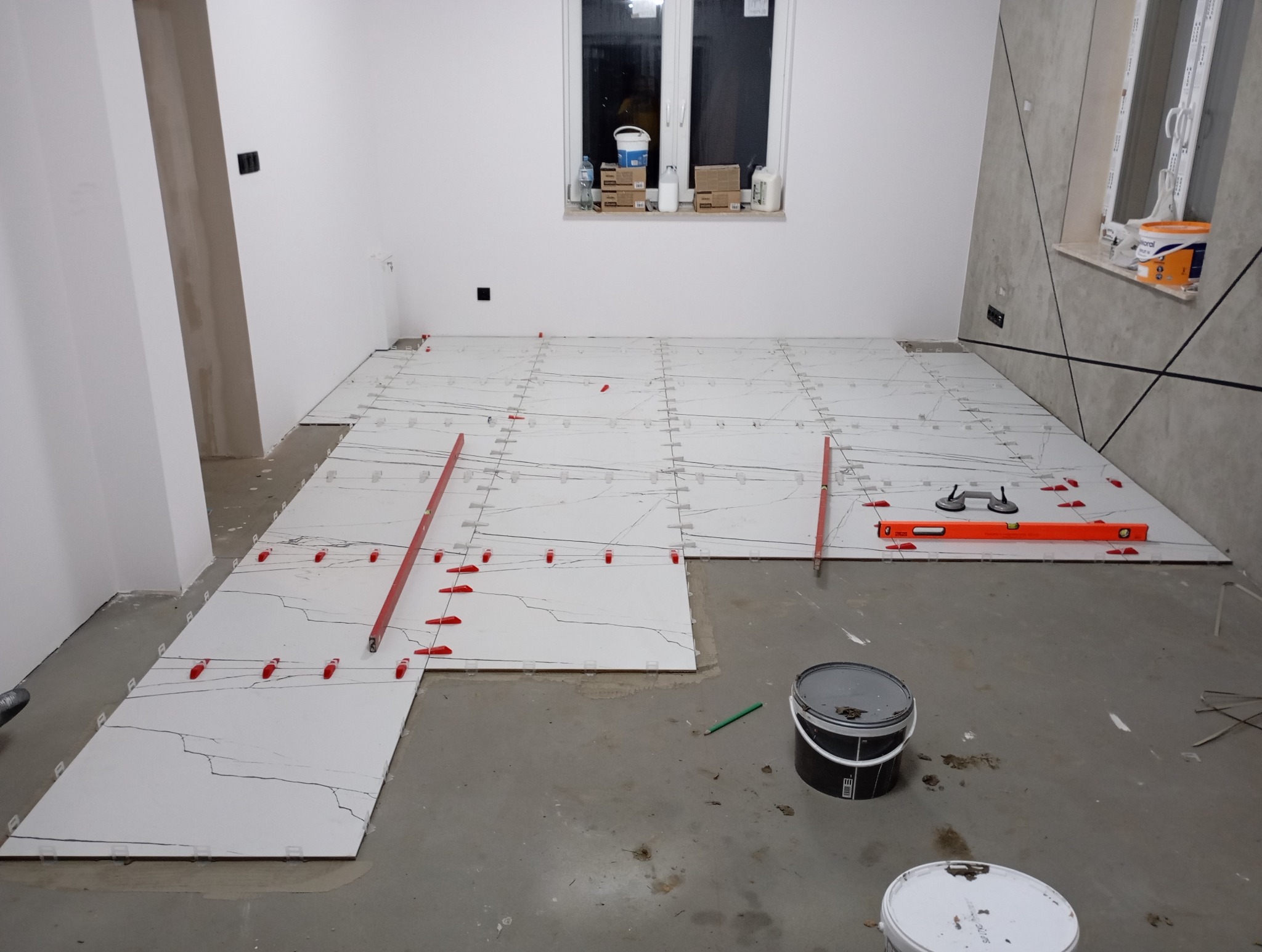 Układanie białych płytek podłogowych z czarnym wzorem żyłkowania. Widoczne poziomice, przyssawki do płytek i czerwone kliny dystansowe. W tle okno i ściana z dekoracyjną fakturą.