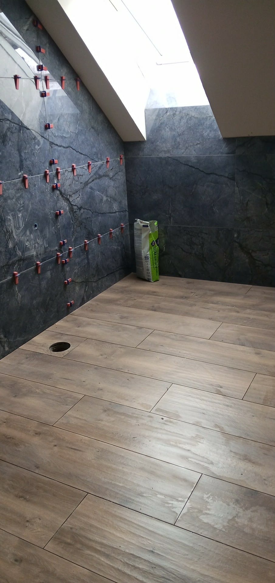 Układanie płytek imitujących drewno na podłodze w łazience ze skośnym sufitem i oknem dachowym. Na ścianach ciemne płytki z czerwonymi krzyżykami dystansowymi.