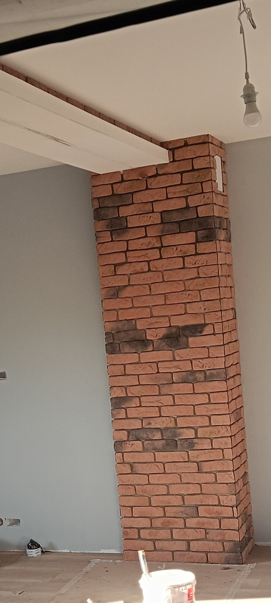 Wykończony fragment ściany z cegły dekoracyjnej w szaro-białym wnętrzu. Widoczny fragment sufitu z lampą. Na podłodze resztki materiałów budowlanych.