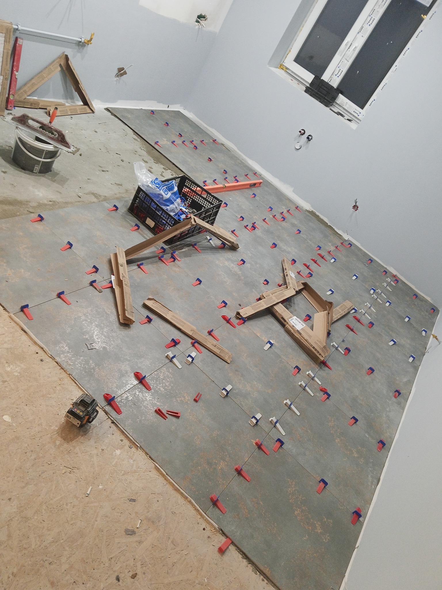 Układanie płytek podłogowych w szarym odcieniu z użyciem czerwonych i białych klipsów poziomujących. Widoczne narzędzia i materiały budowlane. Ujęcie z góry, ukośne.