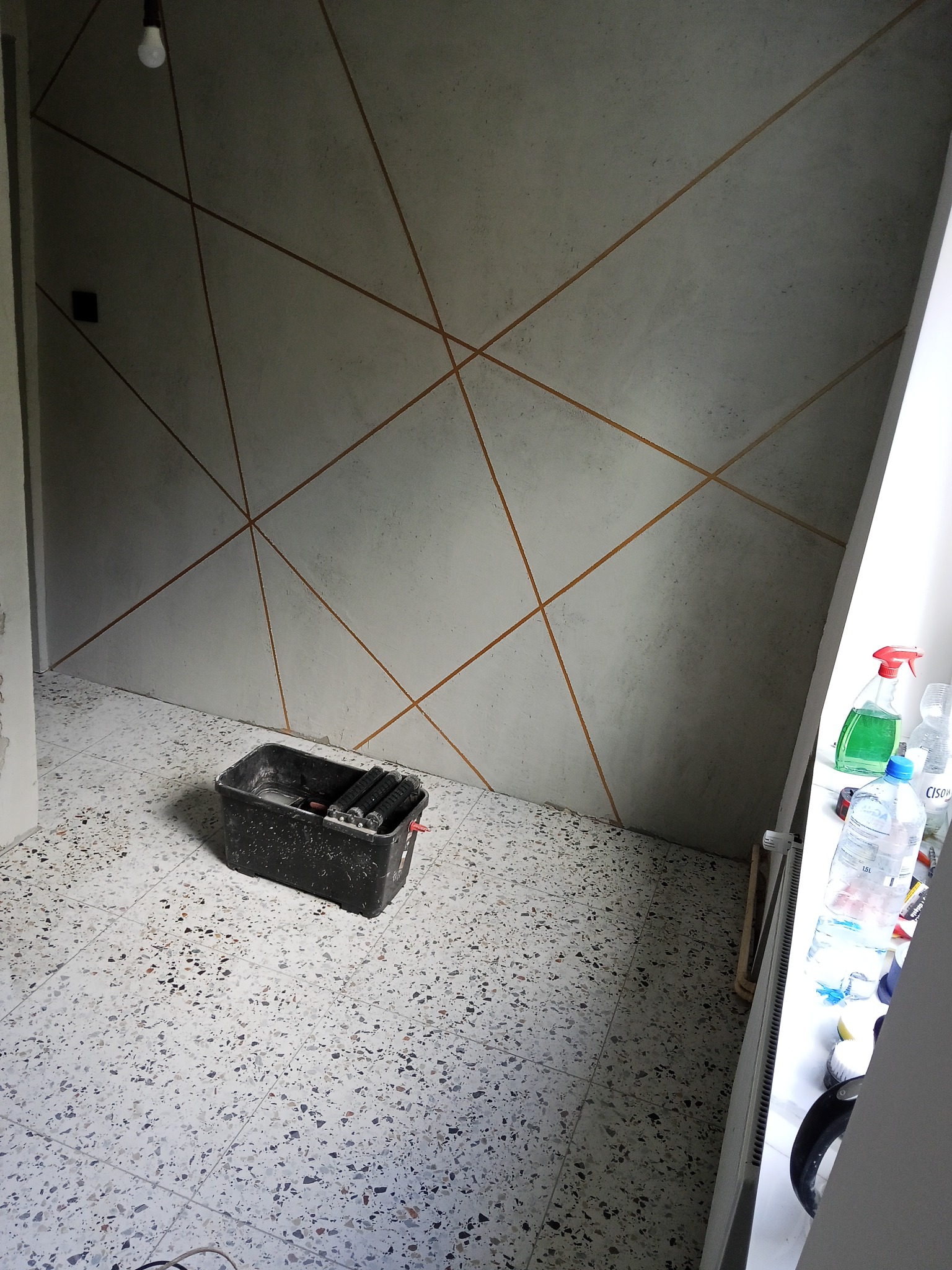 Ściana z geometrycznym wzorem w odcieniach szarości i złota, widoczny kuwet z narzędziami malarskimi, podłoga w terazzo. Wnętrze w trakcie prac wykończeniowych.