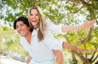 Uśmiechnięty mężczyzna niosący na plecach uśmiechniętą kobietę w plenerze, oboje w białych ubraniach, z rozłożonymi ramionami kobiety, na tle zieleni drzew.