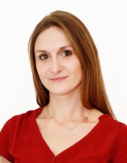 Portret kobiety z długimi, brązowymi włosami, ubranej w czerwoną bluzkę, na białym tle. Widoczny delikatny uśmiech i subtelny makijaż.