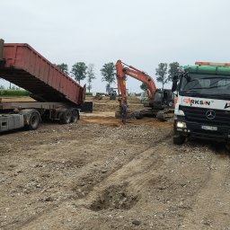 Przedsiębiorstwo Robót Drogowych ARKSAN Arkadiusz Sandrykowicz - Praca ziemne z użyciem koparki i wywrotki na placu budowy. W tle widoczny budynek w trakcie wznoszenia. Na pierwszym planie gruz i piasek.