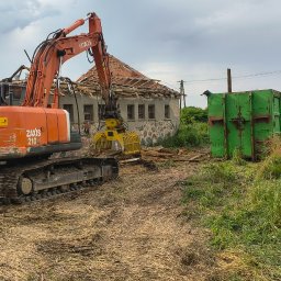 Przedsiębiorstwo Robót Drogowych ARKSAN Arkadiusz Sandrykowicz - Pomarańczowa koparka Hitachi Zaxis 210LC z chwytakiem rozbiera stary budynek z kamiennym cokołem i częściowo zburzonym dachem, obok zielony kontener na gruz.