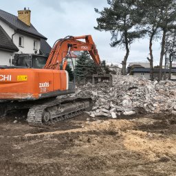 Przedsiębiorstwo Robót Drogowych ARKSAN Arkadiusz Sandrykowicz - Pomarańczowa koparka Hitachi Zaxis 210LC na błotnistym placu budowy, obok hałdy gruzu z betonem i zbrojeniem, z domem i sosnami w tle.
