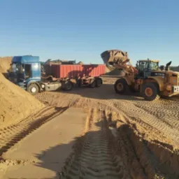 Żółta ładowarka Hyundai HL770-7A nabiera materiał do czerwonej naczepy ciężarówki na placu składowania, w tle widać niebieską kabinę ciągnika siodłowego oraz hałdy materiału.