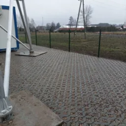 Szara ażurowa nawierzchnia z wypełnieniem żwirowym, obok podstawa konstrukcji wsporczej, w tle ogrodzenie z siatki i słup energetyczny w pochmurny dzień.