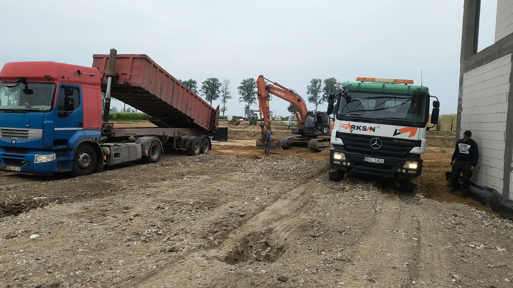 Praca ziemne z użyciem koparki i wywrotki na placu budowy. W tle widoczny budynek w trakcie wznoszenia. Na pierwszym planie gruz i piasek.
