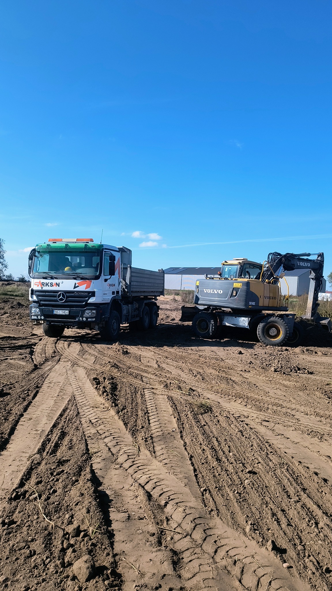 Ciężarówka i koparka Volvo na placu budowy, widoczne ślady opon w ziemi, błękitne niebo. Sprzęt budowlany gotowy do pracy.
