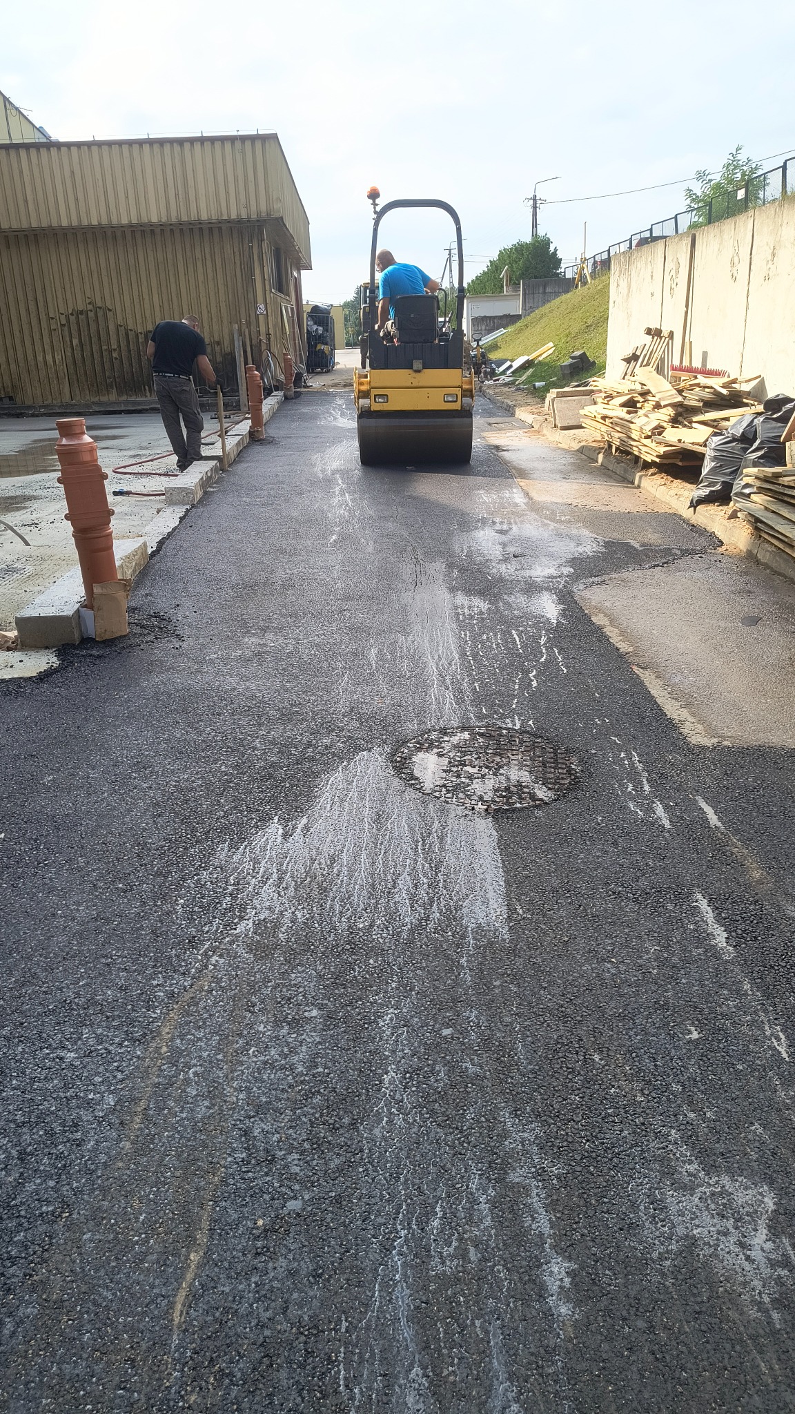 Prace drogowe: walec ubija świeżo wylany asfalt na drodze. Widoczny pracownik wyrównujący krawędzie. Asfalt mokry, z widocznym włazem kanalizacyjnym.