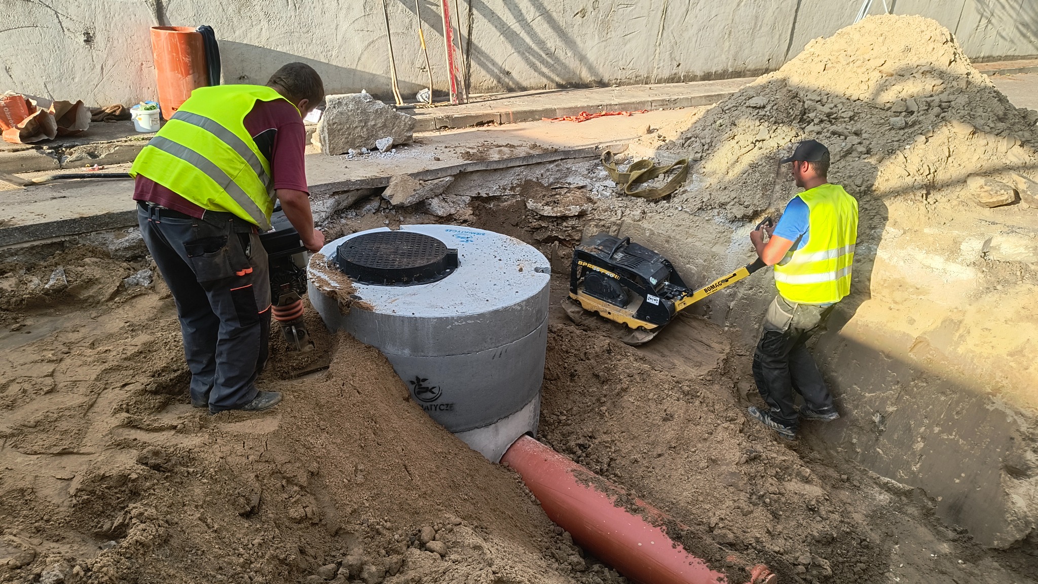 Pracownicy w żółtych kamizelkach zagęszczają grunt wokół betonowego zbiornika z rurą, używając zagęszczarki skokowej i ubijaka w wykopie.
