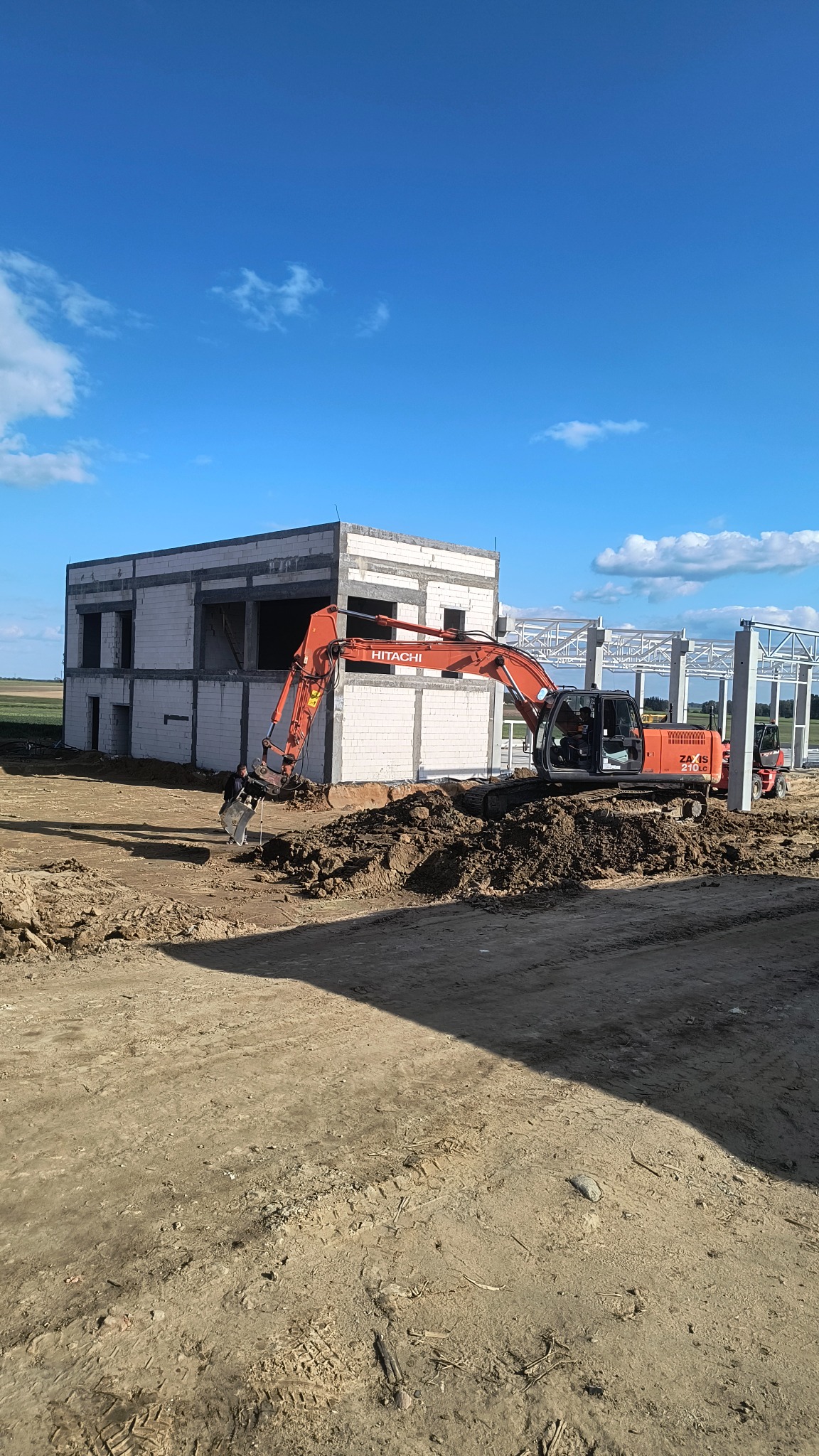 Czerwony Hitachi kopie ziemię przy budowie obiektu w Siemiatyczach. Widoczne ślady opon i fragmenty konstrukcji stalowej. Błękitne niebo z chmurami w tle.