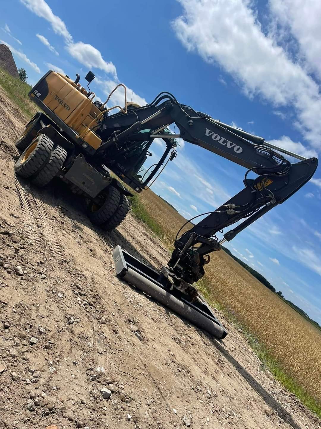 Żółto-czarna koparka Volvo na nierównym, piaszczystym terenie z polem w tle pod błękitnym niebem z chmurami.