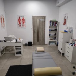 Fizjoterapia Amadeusz Dobies - Gabinet z szarym stołem do masażu, plakaty anatomiczne na ścianach, biurko z laptopem i białe meble. Widoczny czarny dywanik i krzesło na kółkach.