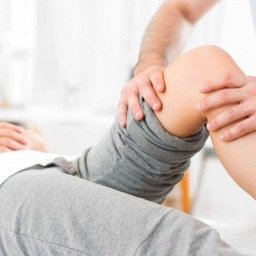 Fizjoterapia Amadeusz Dobies - Pacjent w Puszczykowie leżący na leżance podczas badania stawu kolanowego przez terapeutę. Widoczna ręka terapeuty uciskająca kolano pacjenta.