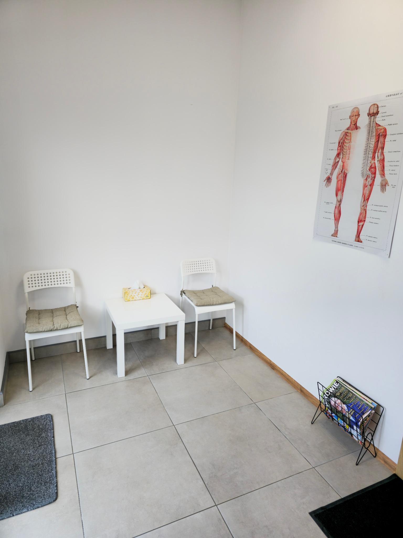 Jasne wnętrze poczekalni gabinetu z dwoma krzesłami z poduszkami, białym stolikiem z pudełkiem chusteczek, plakatem anatomicznym i stojakiem na czasopisma.