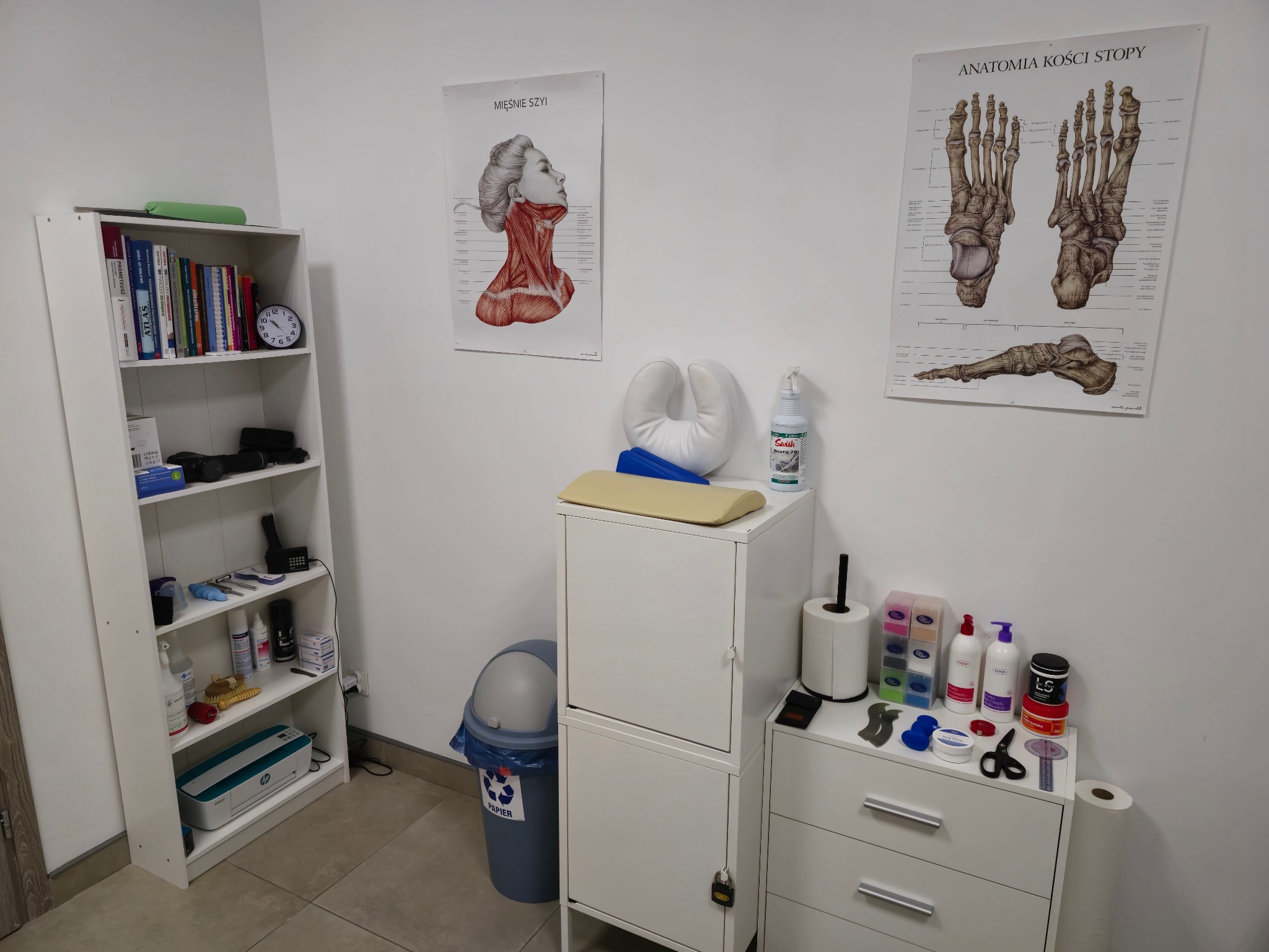 Gabinet z plakatem mięśni szyi i anatomii kości stopy, białe meble, książki, kosmetyki i akcesoria do zabiegów.