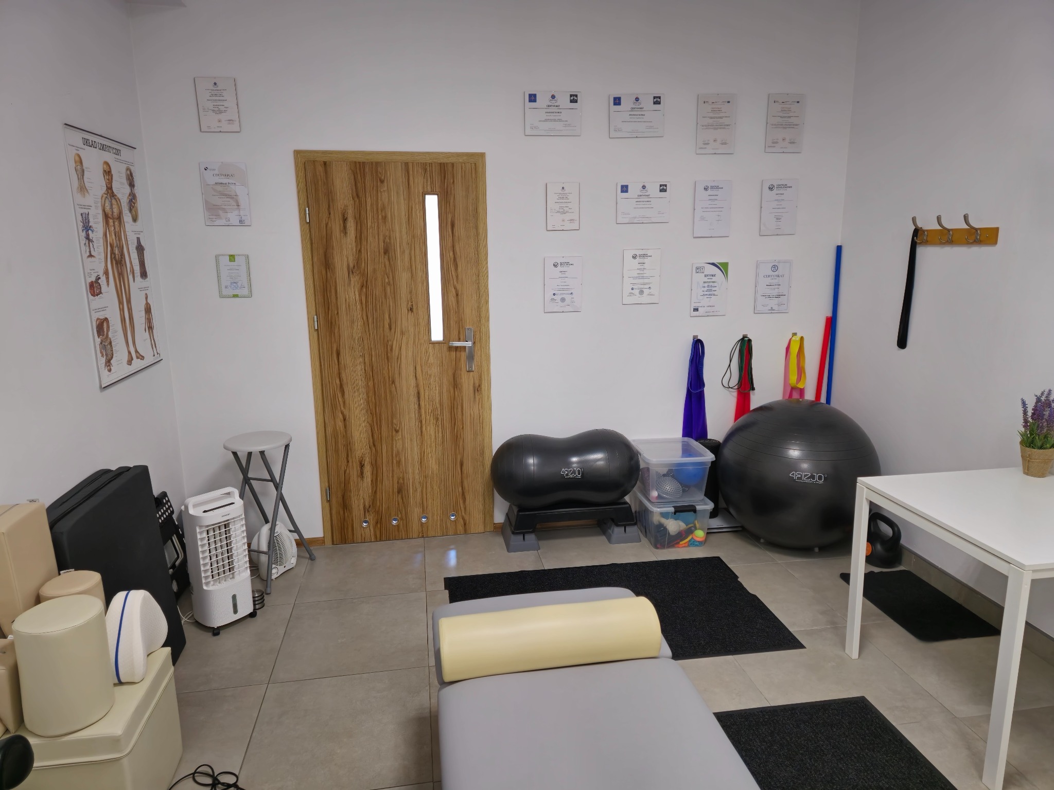 Gabinet fizjoterapii z certyfikatami na ścianie, piłkami do ćwiczeń, leżanką z wałkiem i plakatem z układem limfatycznym.