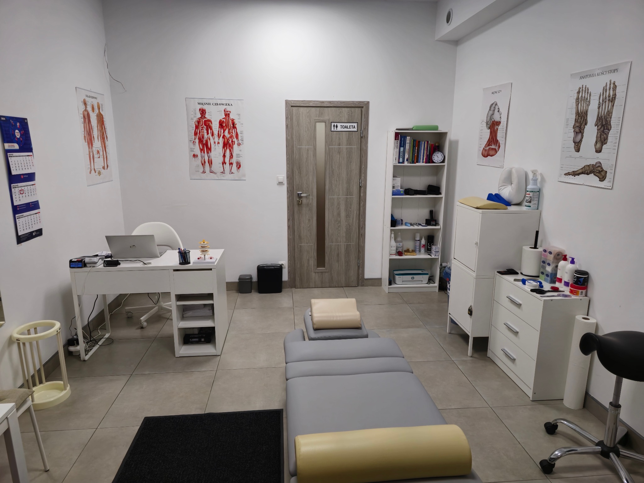 Gabinet z szarym stołem do masażu, plakaty anatomiczne na ścianach, biurko z laptopem i białe meble. Widoczny czarny dywanik i krzesło na kółkach.