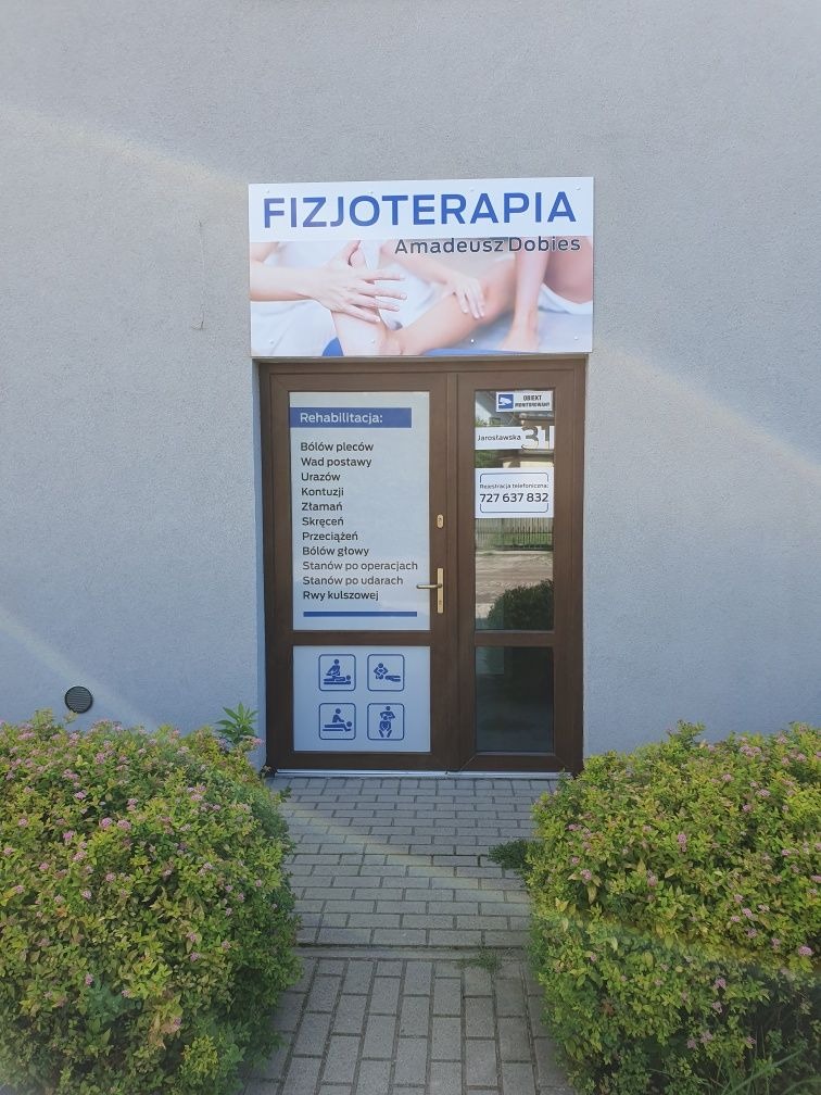 Wejście do gabinetu z szyldem 'Fizjoterapia Amadeusz Dobies', na drzwiach wykaz usług rehabilitacyjnych oraz piktogramy i numer telefonu, przed wejściem krzewy.