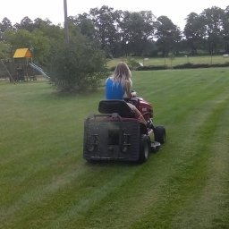 Green Garden Tomasz Szypulski - Kobieta kosi trawnik traktorkiem, widok z tyłu na równo przystrzyżoną trawę z pasami, plac zabaw w tle.