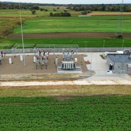 PG-COMPLEX - Widok z lotu ptaka na ogrodzony teren stacji transformatorowej z budynkiem technicznym, urządzeniami elektrycznymi i słupami wysokiego napięcia na tle zielonych pól uprawnych i odległych zabudowań.