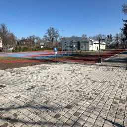 PG-COMPLEX - Nowo ułożona, szara kostka brukowa na terenie rekreacyjnym z boiskiem i halą sportową w tle, w słoneczny dzień. Widoczny znak parkingu.