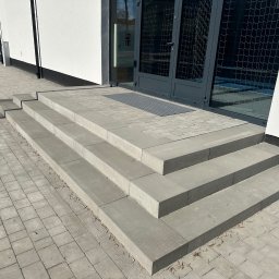 PG-COMPLEX - Schody wejściowe z szarej kostki brukowej prowadzące do budynku z szarymi drzwiami. Kostka ułożona w regularny wzór, widoczny fragment chodnika i elewacji budynku.