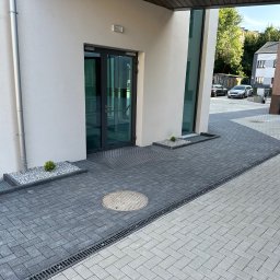 PG-COMPLEX - Ułożona, dwukolorowa kostka brukowa przed budynkiem z szarymi oknami i drzwiami. Widoczny właz kanalizacyjny i odpływ liniowy. Małe rabaty z roślinami.