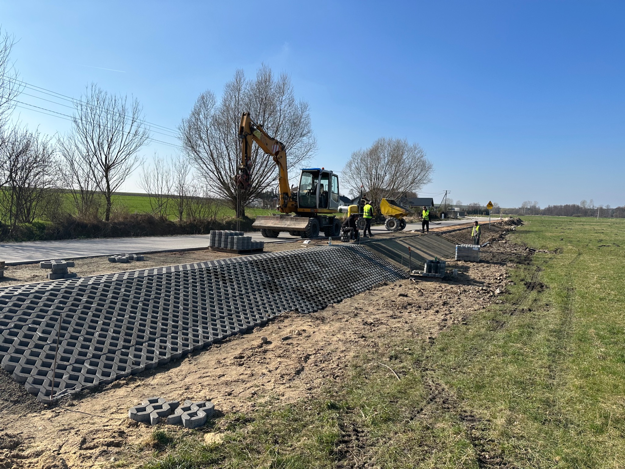 Żółta koparka i pracownicy w kamizelkach odblaskowych przy budowie drogi z ażurowych płyt betonowych, widoczne ułożone płyty i przygotowany teren, w tle drzewa bez liści i błękitne niebo.