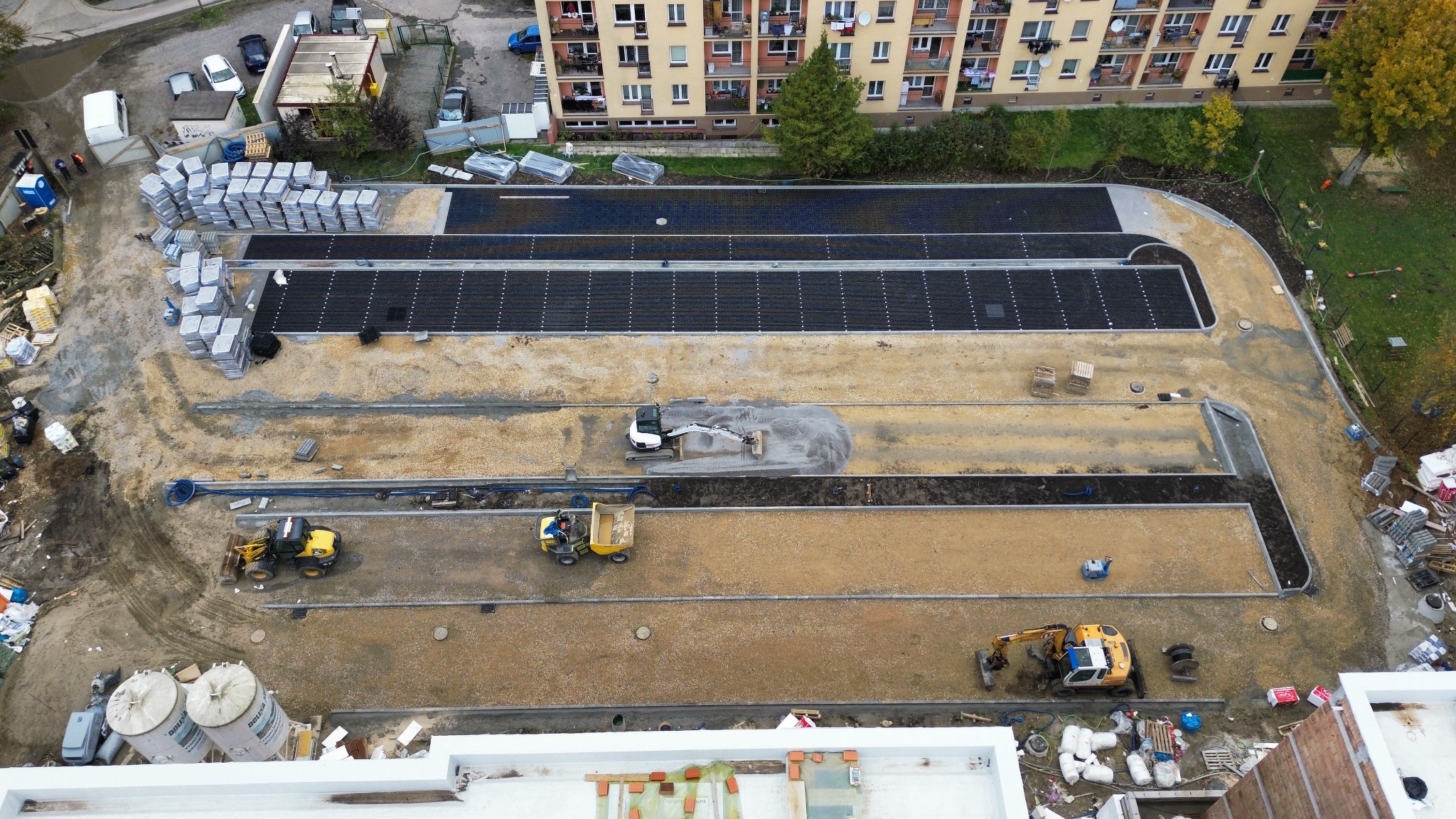 Widok z góry na plac budowy z kilkoma rzędami przygotowywanej nawierzchni, gdzie w górnych rzędach ułożona jest czarna kostka, a w dolnych trwa rozprowadzanie piasku przez maszyny budowlane,...