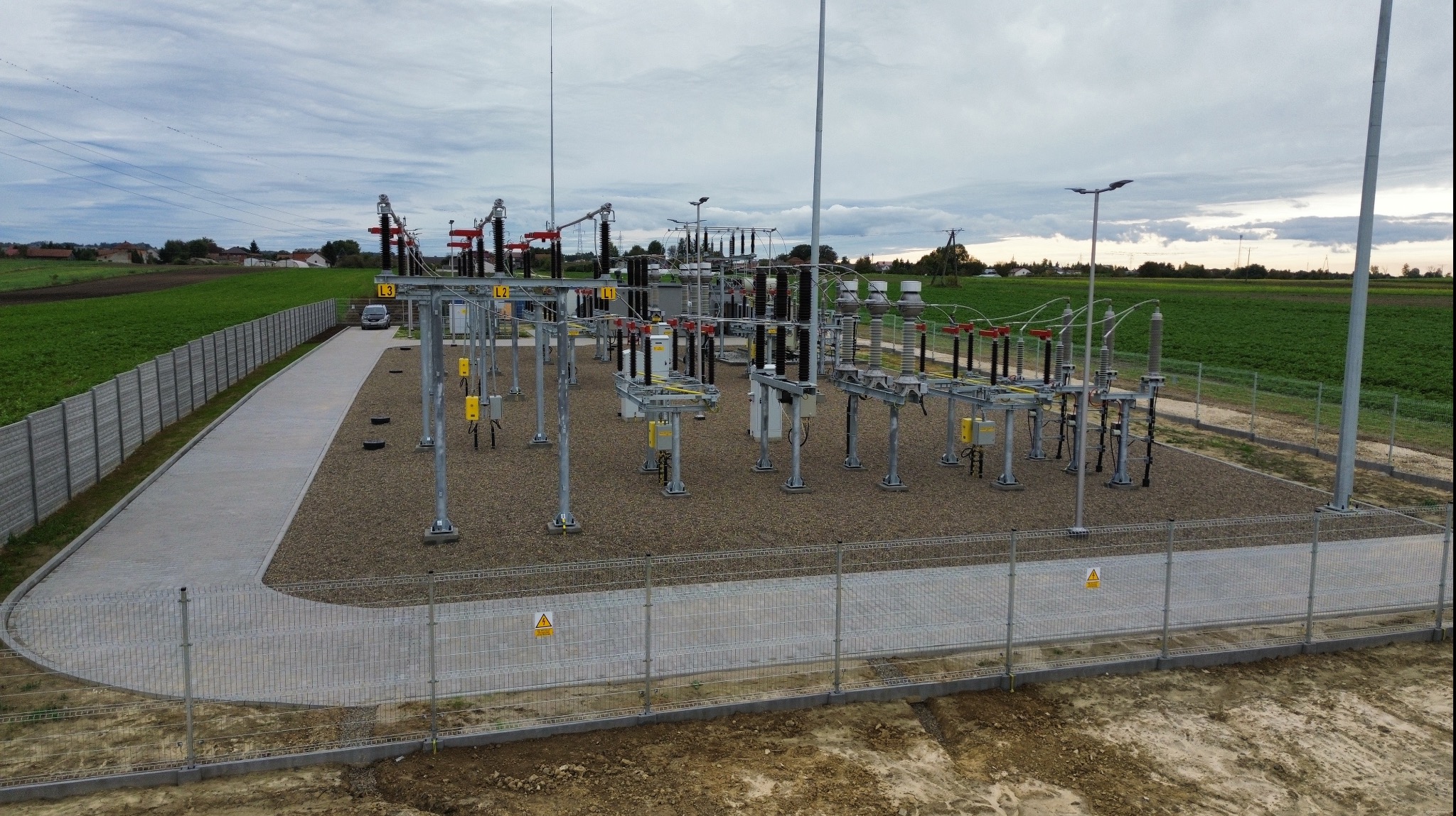 Widok z góry na ogrodzony teren stacji transformatorowej z nową nawierzchnią z kostki brukowej i utwardzonym placem, otoczoną zielonymi polami pod pochmurnym niebem.