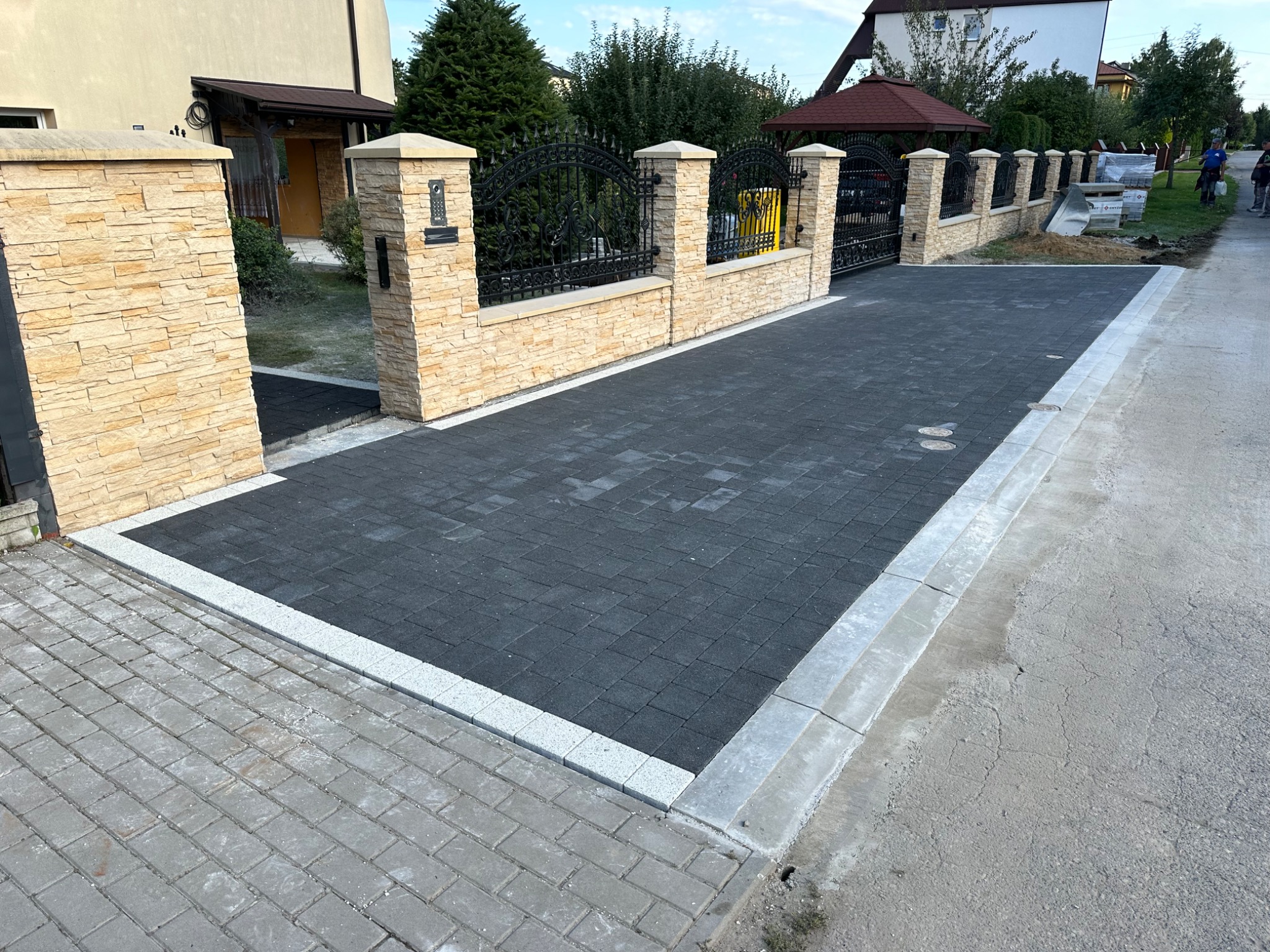 Nowoczesny podjazd z ciemnej kostki brukowej, obrzeża z jasnej kostki. Ogrodzenie z elementami kamiennymi i kutymi, brama wjazdowa. Widok z zewnątrz posesji.
