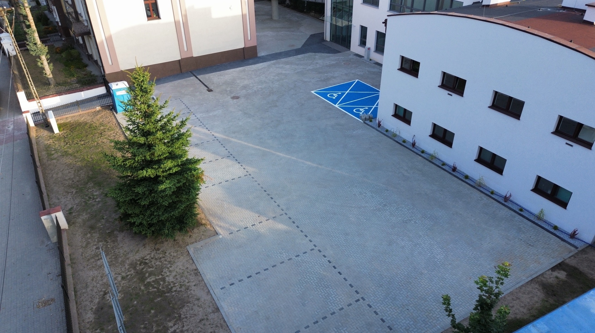 Z lotu ptaka: parking z kostki brukowej z wyznaczonym miejscem dla niepełnosprawnych, obok budynki i drzewo. Widoczne detale ułożenia kostki.