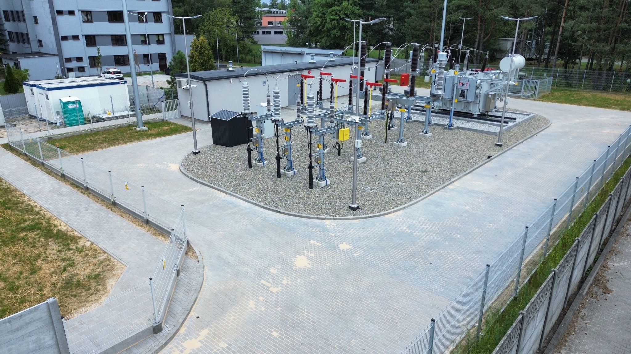 Zabezpieczony teren stacji transformatorowej z utwardzonym placem z kostki brukowej i ogrodzeniem panelowym. Widok z góry ukazuje detale infrastruktury.