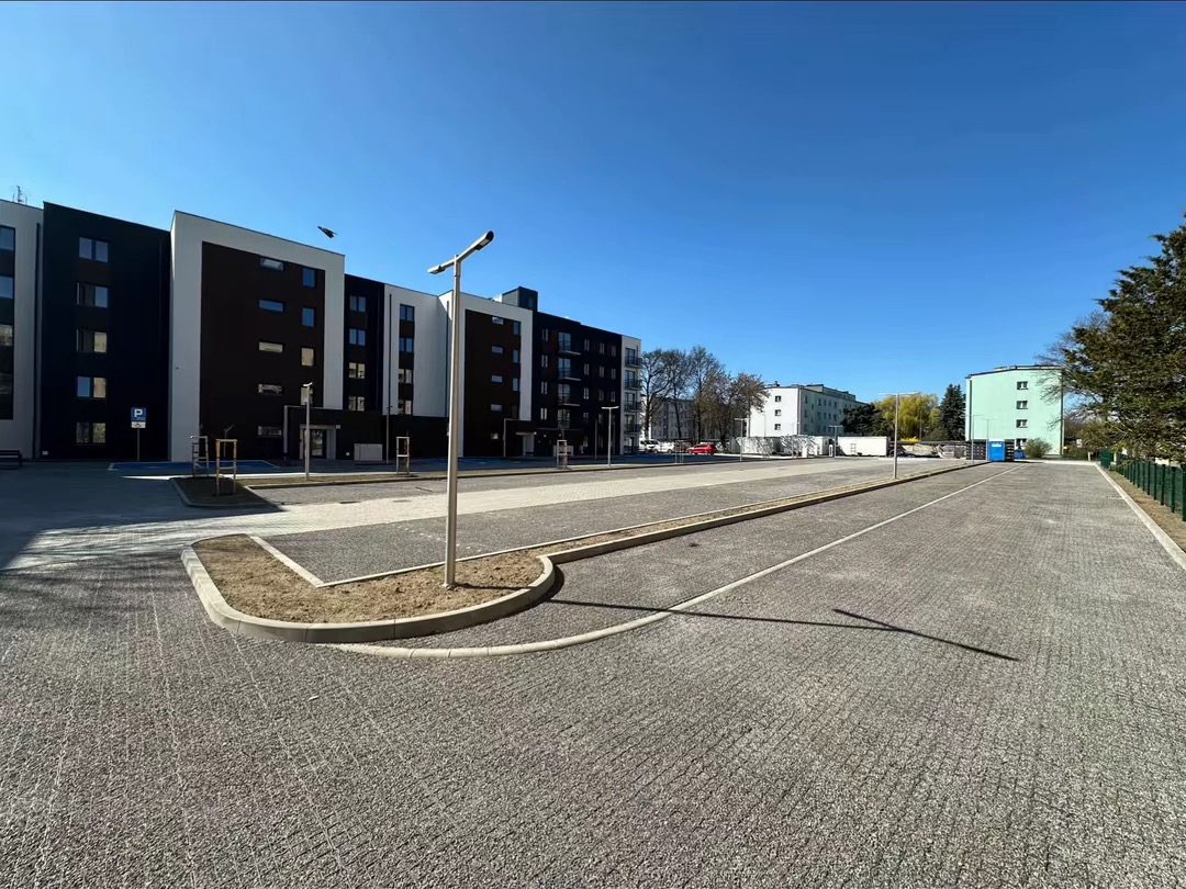 Nowoczesny parking z kostki brukowej przy budynku, z oświetleniem i wyznaczonymi miejscami, w słoneczny dzień. Widok na budynek i drzewa w tle.