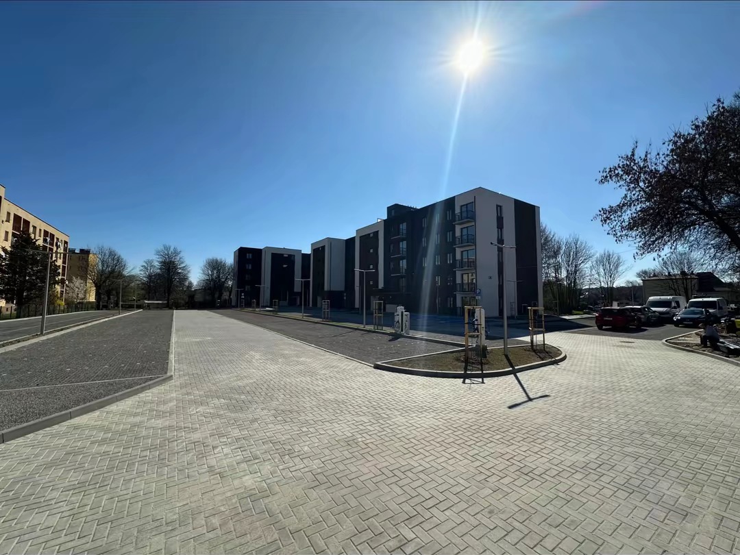 Nowoczesne osiedle z utwardzonym terenem z kostki brukowej w jodełkę, oświetlone słońcem, z widocznymi budynkami mieszkalnymi i zaparkowanymi samochodami.