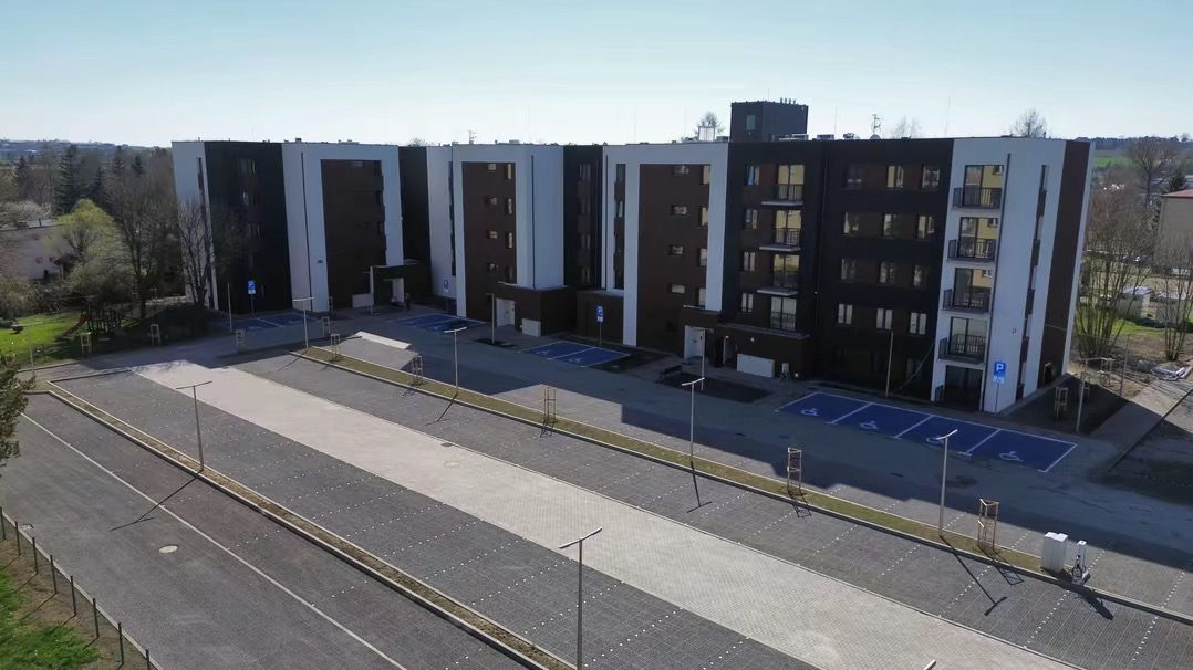 Nowoczesny budynek wielorodzinny z dużym parkingiem z kostki brukowej. Widoczne miejsca parkingowe dla niepełnosprawnych i oświetlenie.